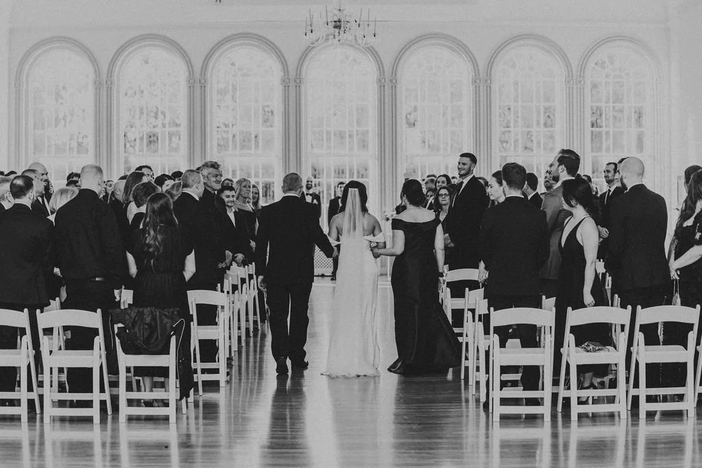 A formal wedding ceremony with guests standing on either side of the aisle with the bride and groom holding hands at the altar in a large, bright hall with tall arched windows.