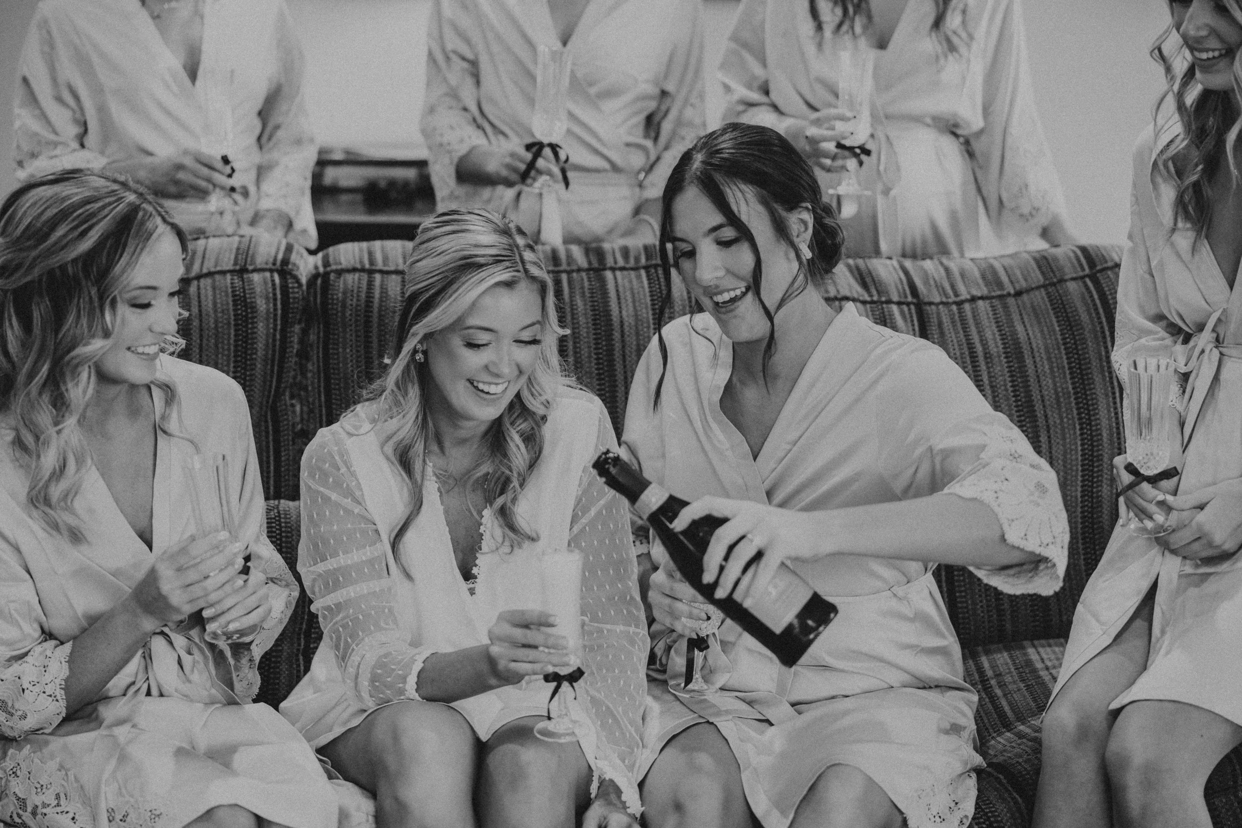 Women in robes enjoying a celebration, some holding glasses and one opening a bottle, in a cozy room.