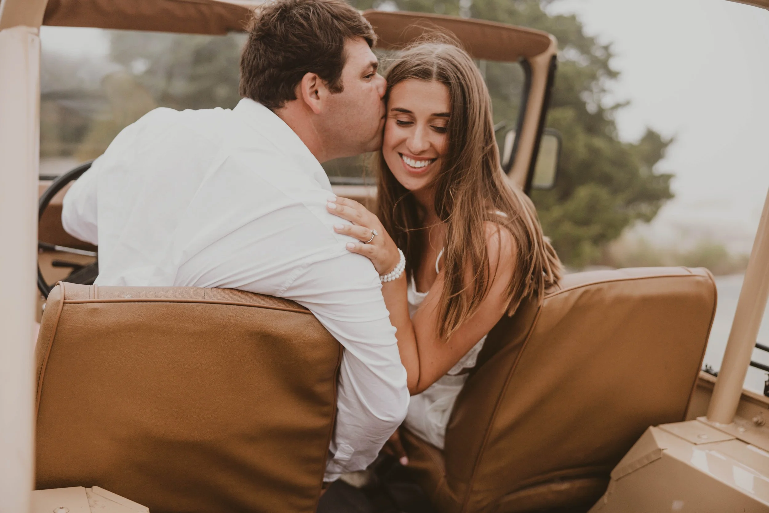 A man kissing a woman on the forehead in a vintage convertible car with open top during daytime, both smiling; the woman has long brown hair and wears a white dress, the man has short brown hair and wears a white shirt.