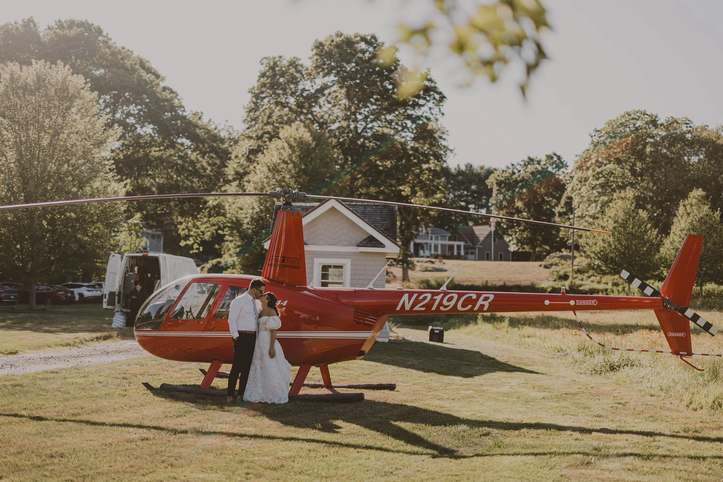 A couple in wedding attire standing close to each other next to a red helicopter parked on a grassy area, with trees and houses in the background.