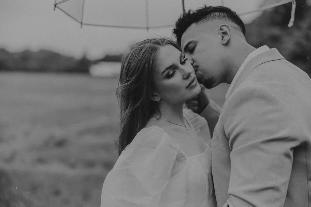 A black and white photo of a couple sharing an intimate moment under a parasol outdoors. The woman has long hair and is wearing a blouse, while the man is dressed in a suit. They are close, with their foreheads touching.