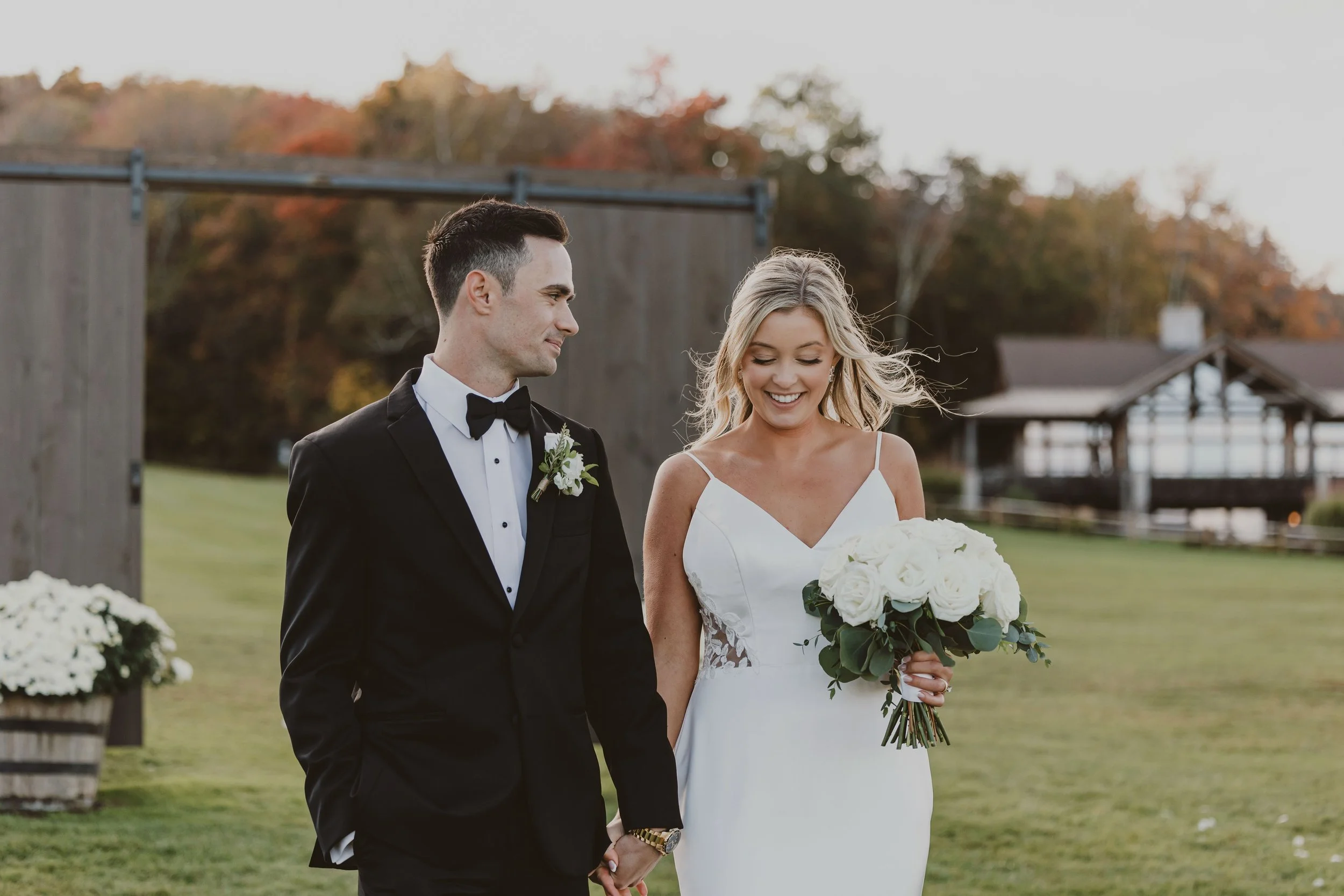 A bride and groom holding hands outdoors on their wedding day, with the bride smiling and holding a bouquet of white roses, and the groom looking at her. They are dressed in formal wedding attire, standing on a grassy field with autumn trees and a wo