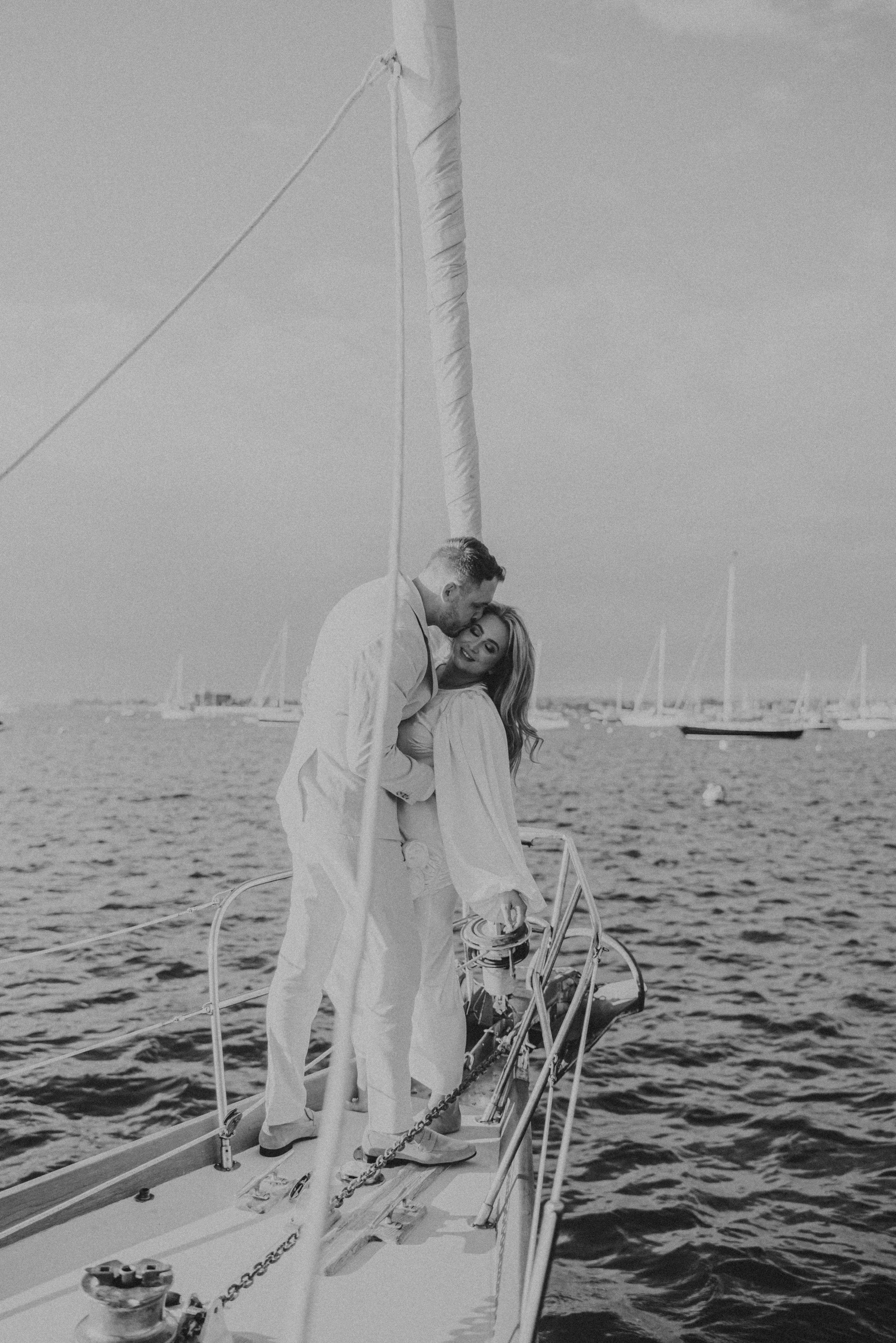 A couple dressed in white on a sailboat, embracing by the water with sailboats in the background.