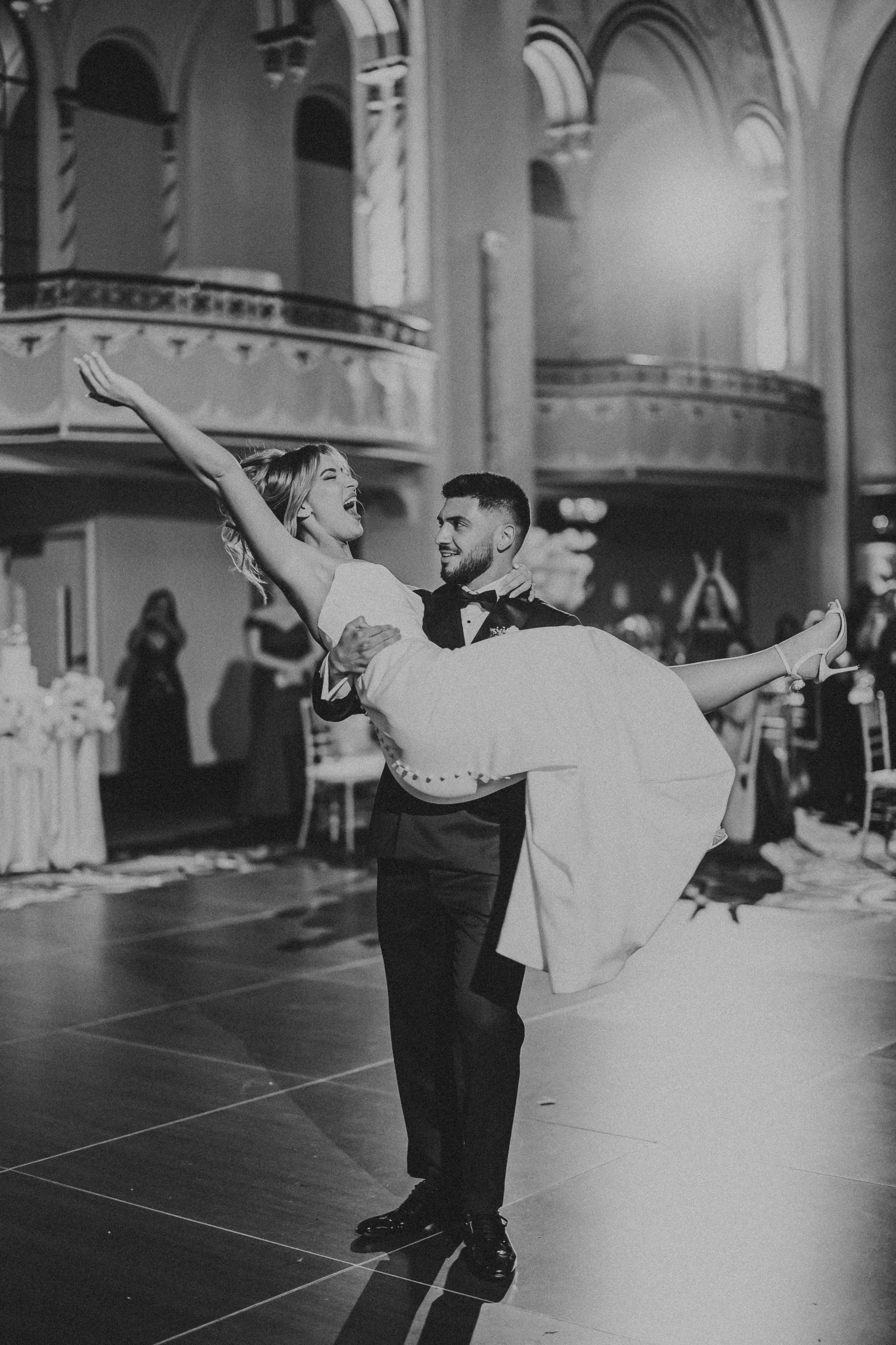 A man in a tuxedo is dancing and holding a woman in a white dress in a ballroom with high arched ceilings and balconies, with guests in the background.