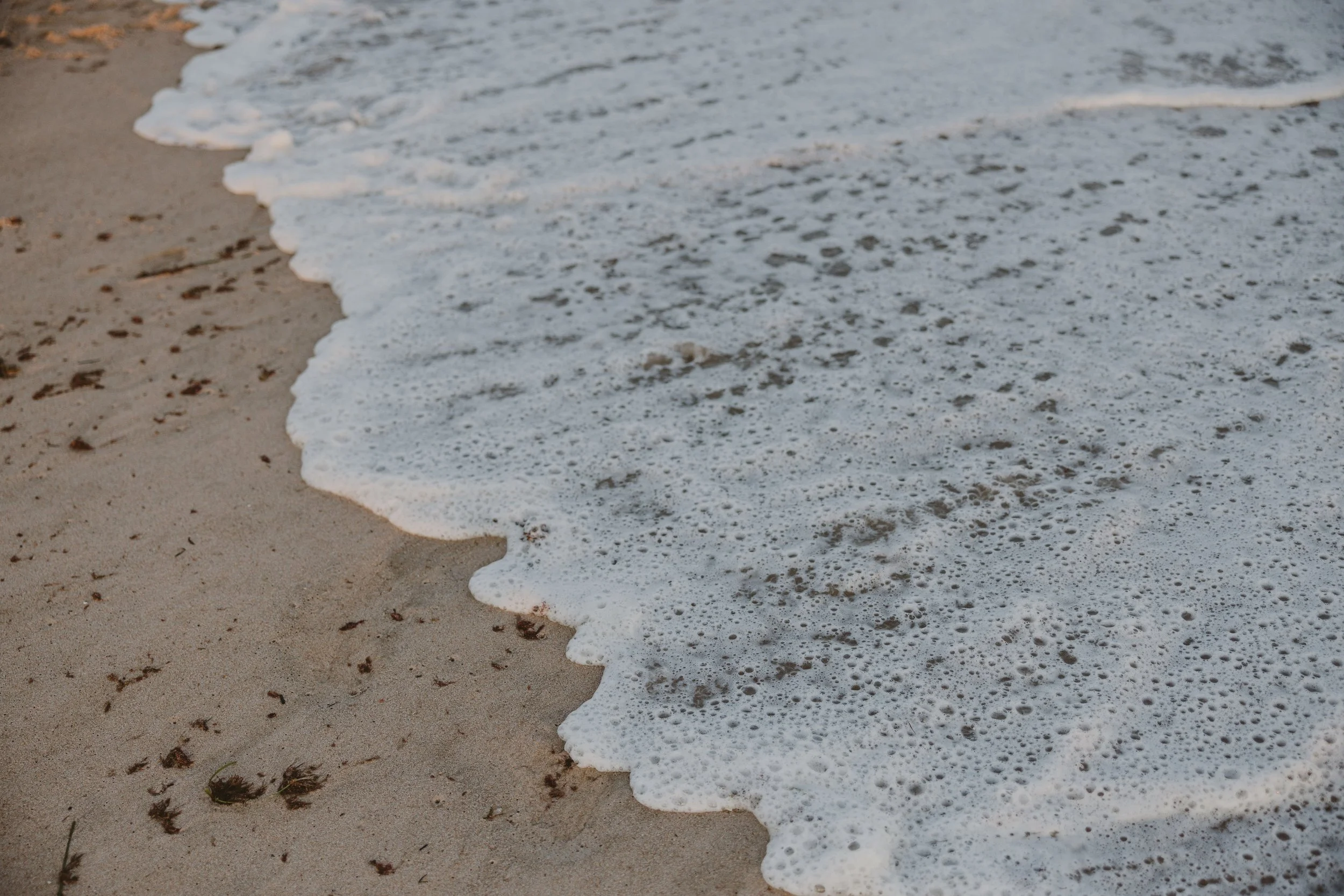 Close-up photo of ocean waves gently washing onto sandy beach with small pebbles and seaweed.