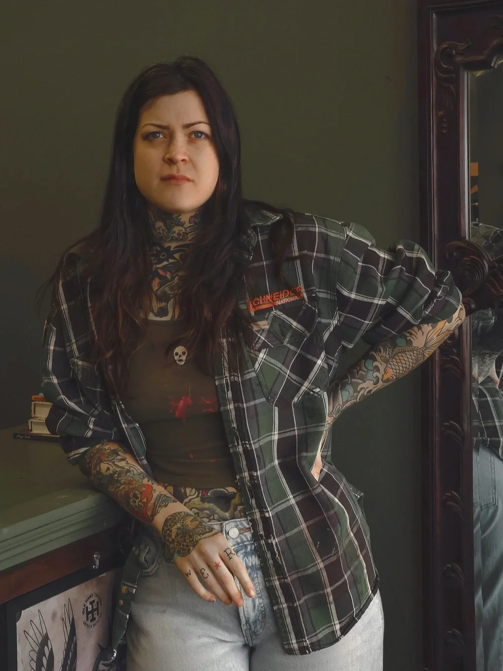 A woman with long dark hair and tattoos on her arm and hand, wearing a black tank top and a green plaid shirt, standing next to a mirror with a dark wood frame. She is looking at the camera with a serious expression and has her hand on her hip.