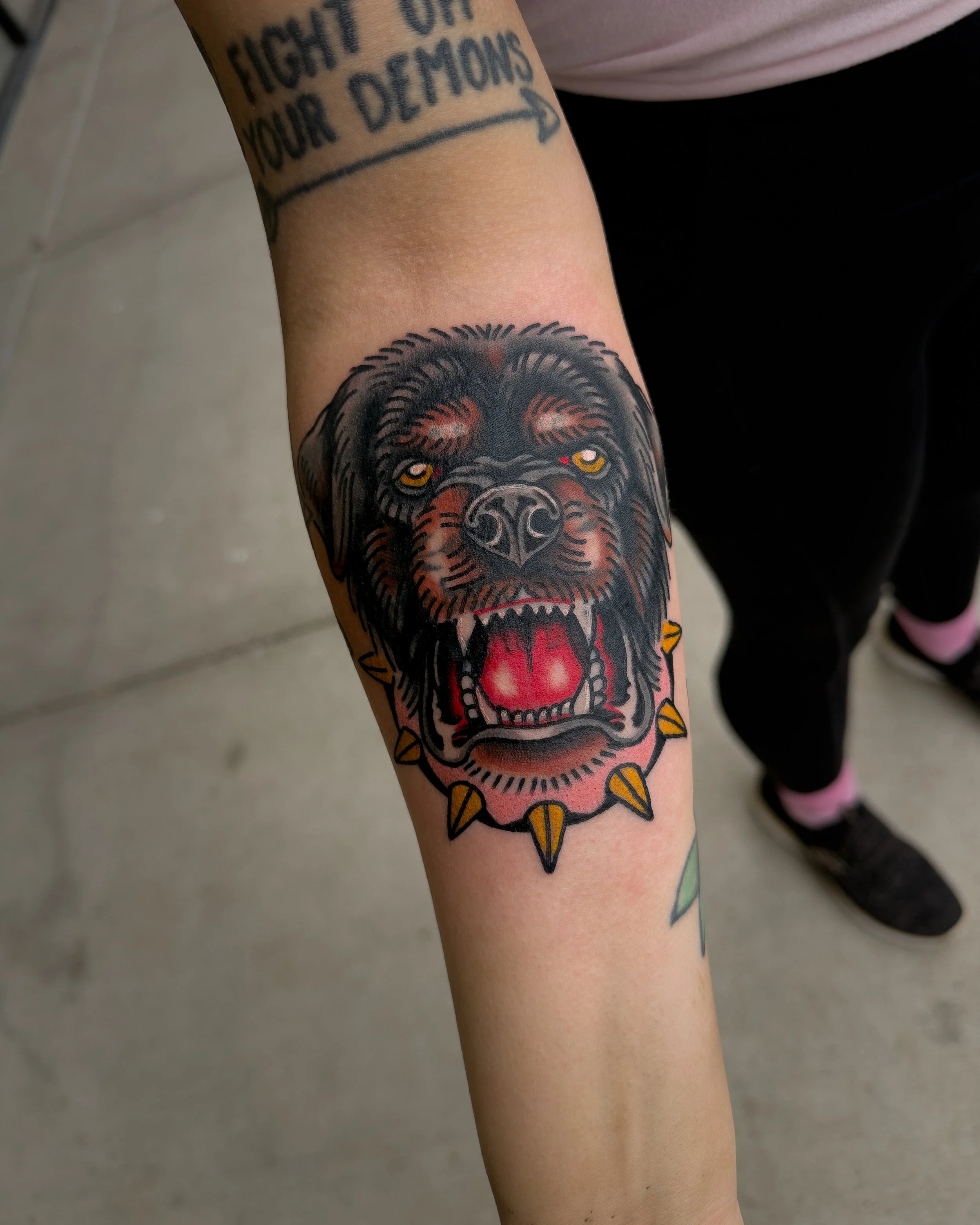 Close-up of a person's arm showing a tattoo of a roaring dog with yellow eyes and an open mouth, surrounded by a spiked border, with part of a tattoo reading 'FIGHT' visible above.