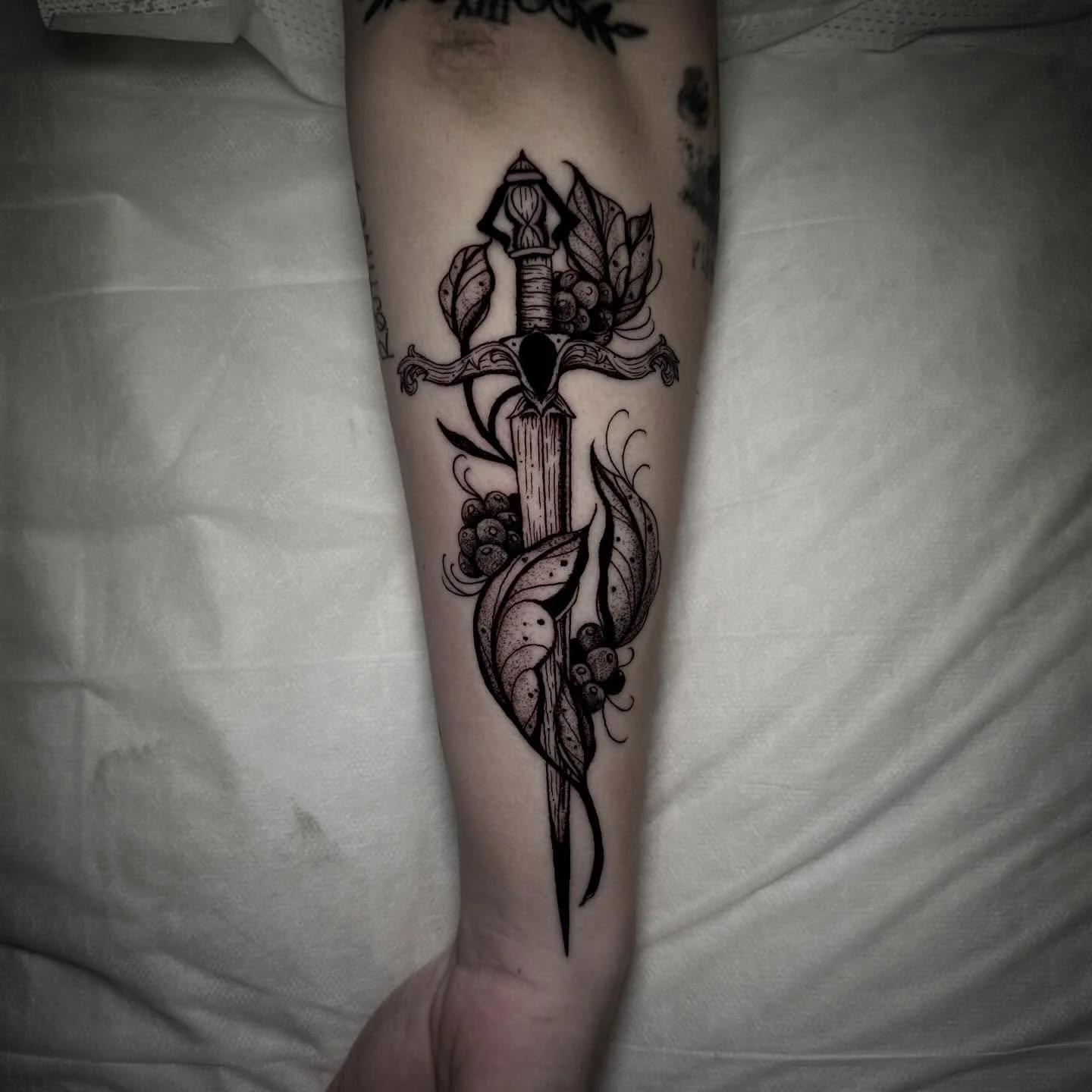 Tattoo of a dagger with a detailed hilt and blade, surrounded by leaves and berries, on a person's forearm.