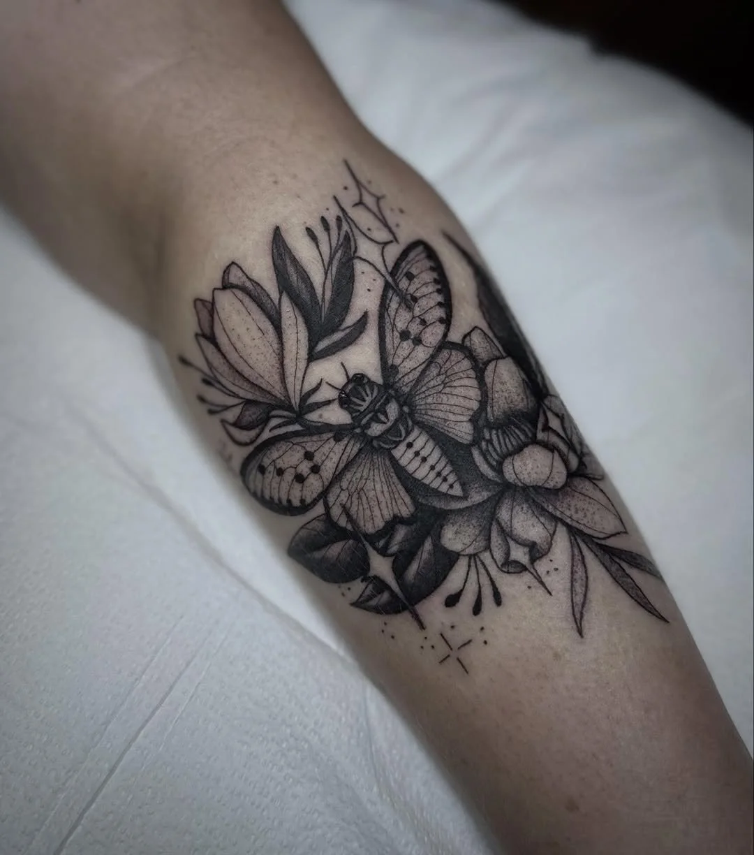 A black and gray tattoo of a butterfly, flowers, leaves, and small decorative elements on a person's arm.