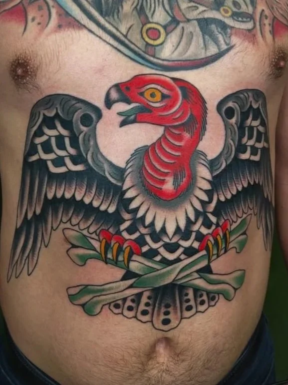 A man with tattoos on his arms, wearing a black T-shirt and a wristwatch, standing indoors with a colorful tiger face artwork behind him. The artwork features a tiger with an open mouth, showing sharp teeth and a green tongue. There are bottles of tattoo ink and tattoo equipment on the counter behind him.