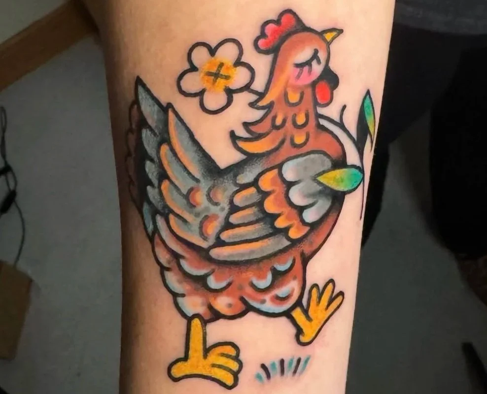 Colorful tattoo of a cartoon-style rooster with a flower and a rainbow on its wing on a person's arm.