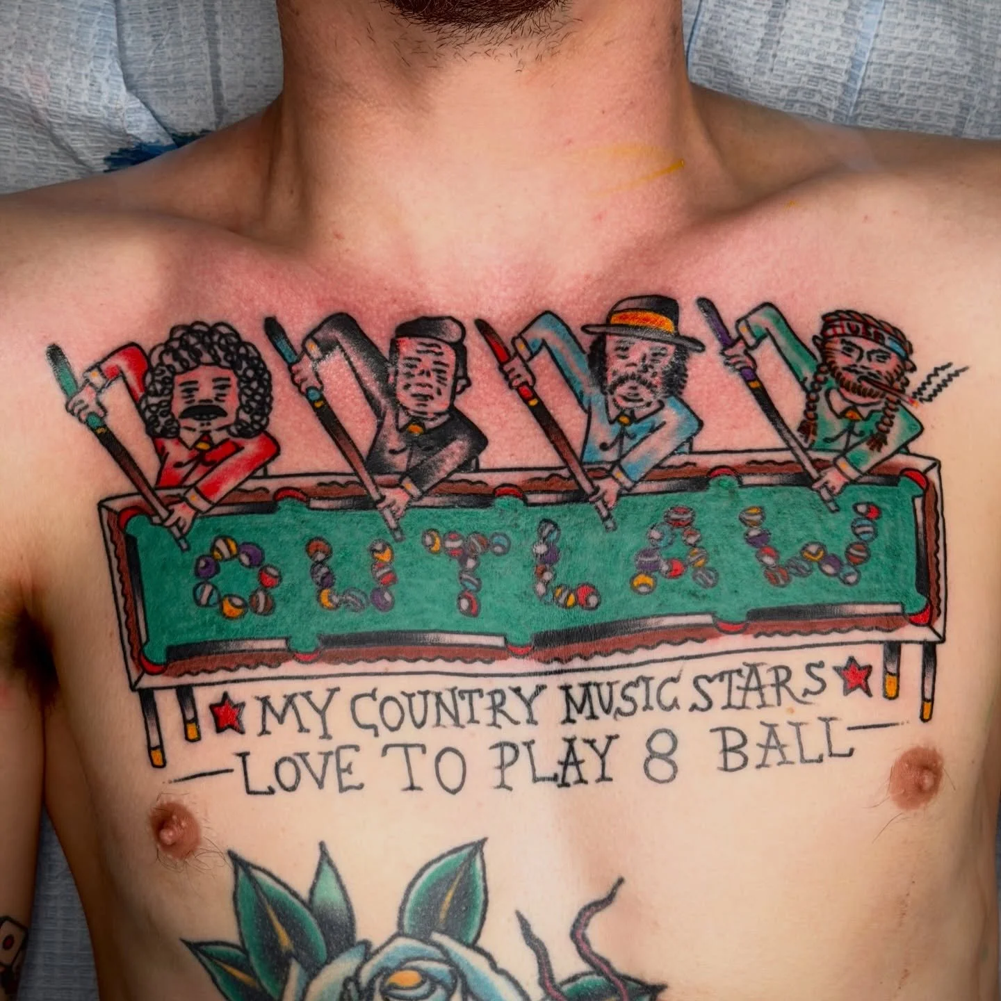 A young man with glasses, wearing a brown baseball cap and a black Harley Davidson sweatshirt, standing in front of a framed tattoo artwork on a green wall. There are tattoo designs on the wall behind him.