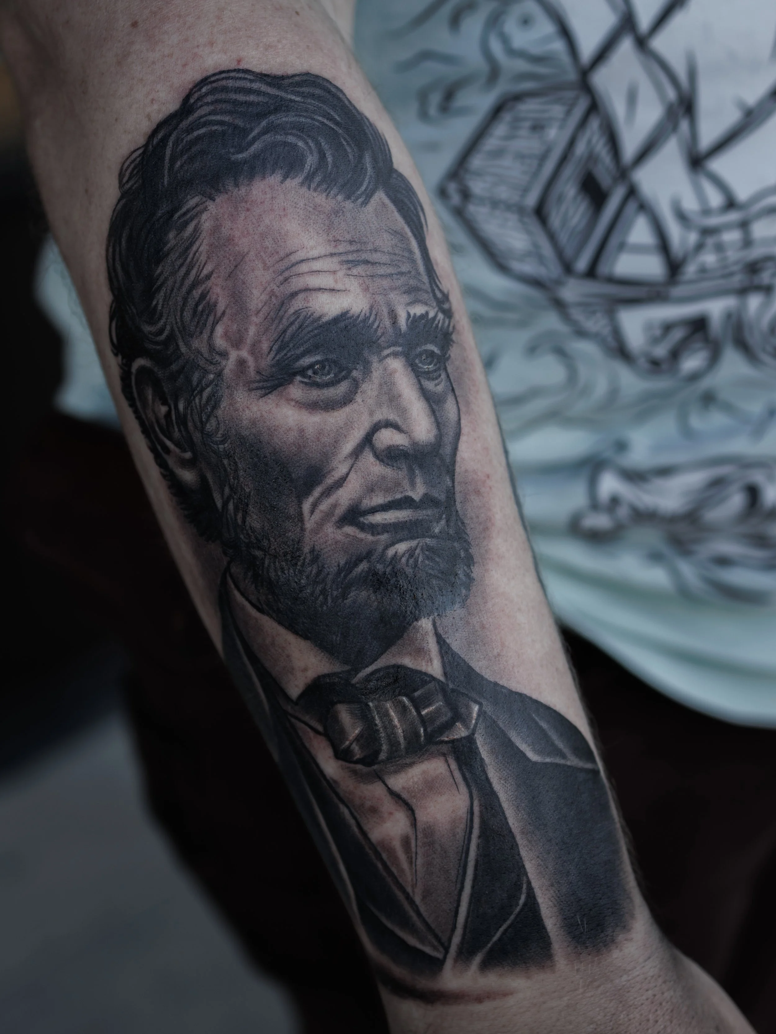 Close-up of a tattoo on a person's arm depicting a realistic black and gray portrait of a distinguished man with slicked-back hair and a bow tie.