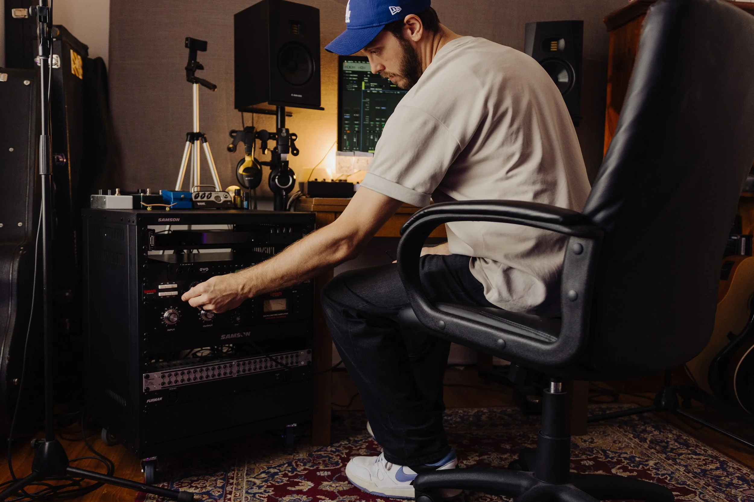 Man wearing a blue cap and beige shirt sitting in a recording studio, adjusting audio equipment on a rack.