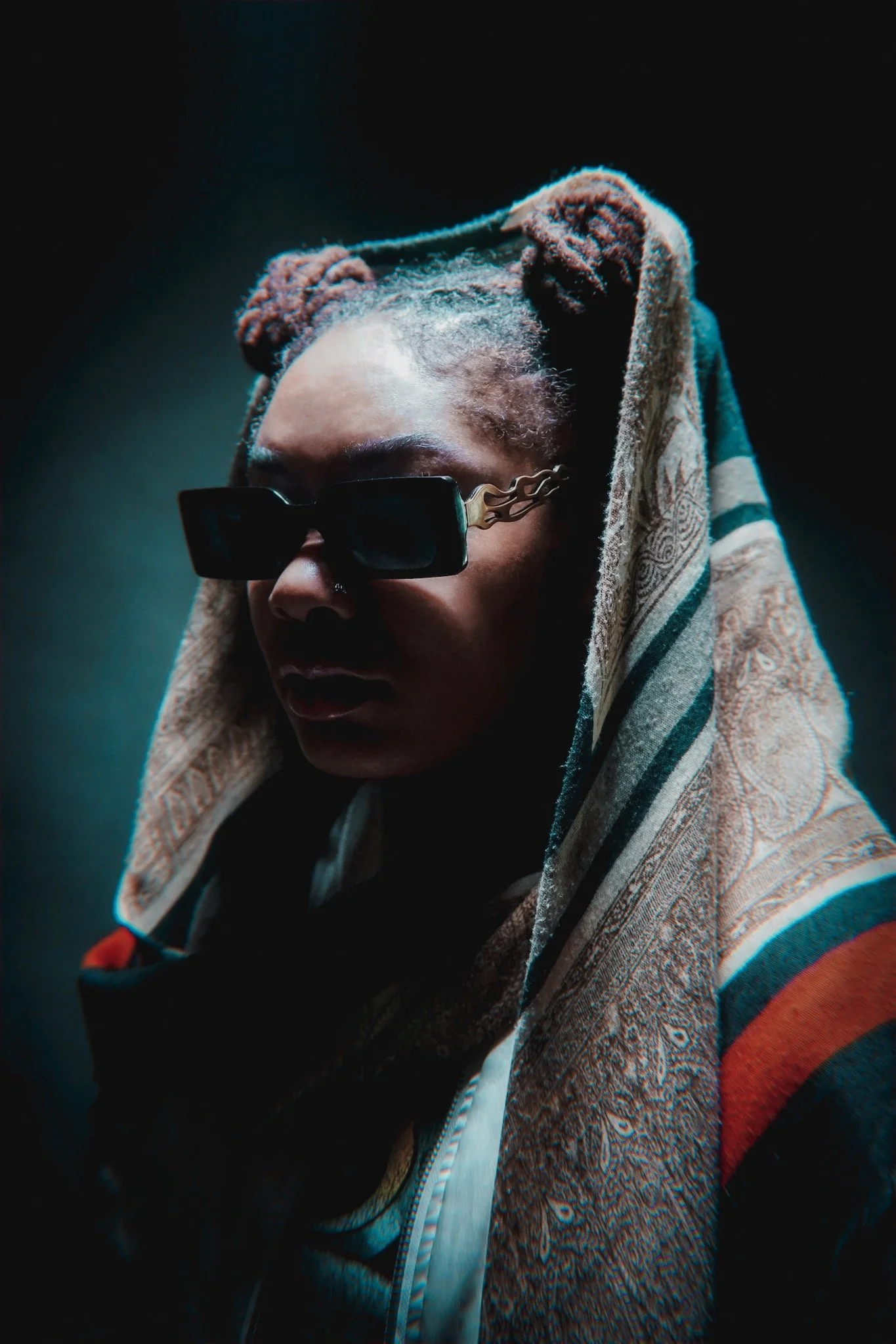 Person wearing sunglasses with a hooded shawl, in a dimly lit setting, casting shadows on the face.