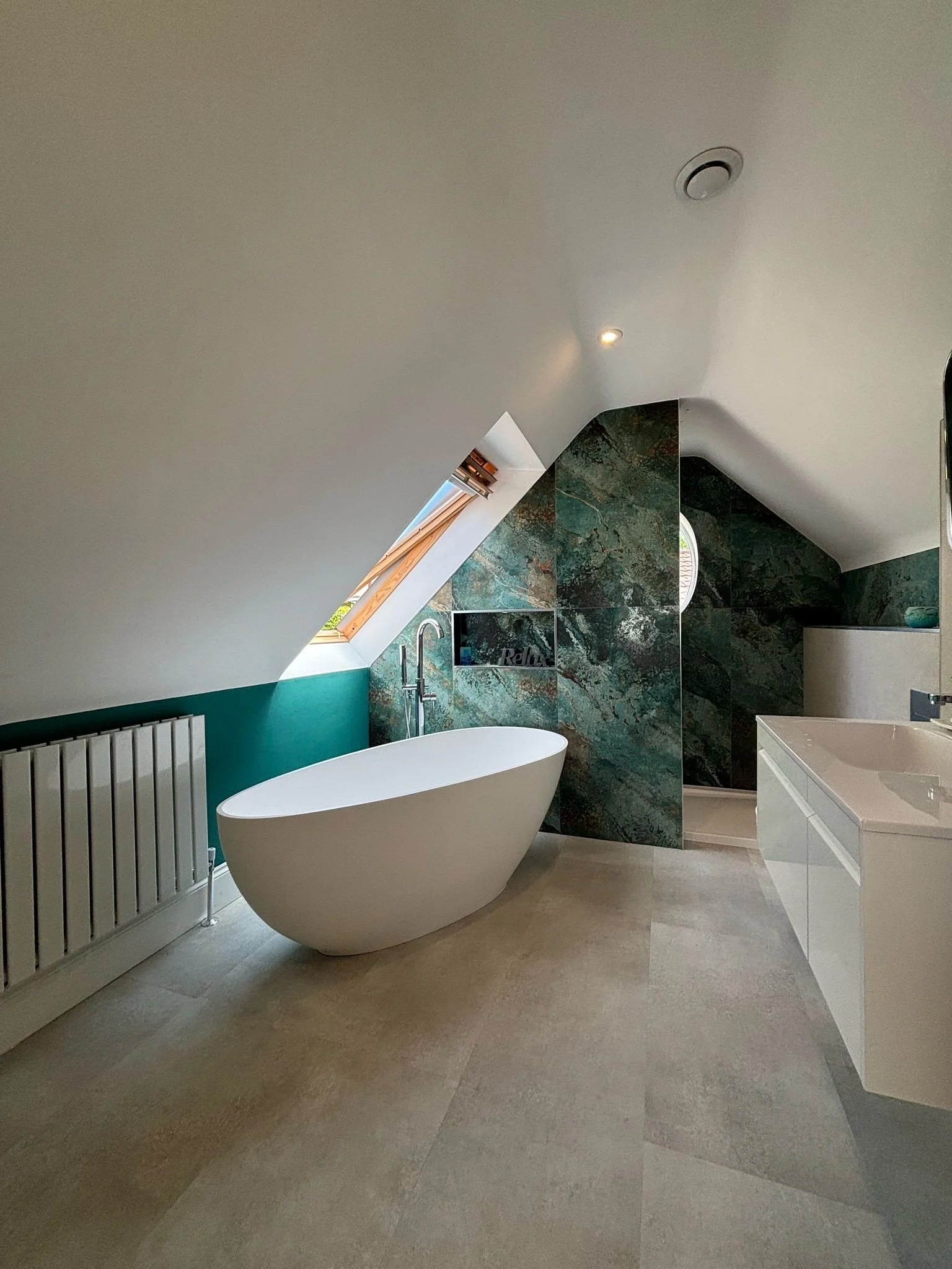 Modern bathroom with a freestanding bathtub, sloped ceiling with skylight, green marble wall accents, white vanity, and beige tile floor.