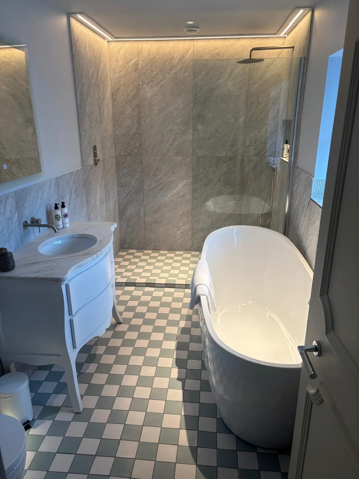 A modern bathroom featuring a freestanding bathtub, a walk-in glass shower with a rain showerhead, a marble vanity with a sink, and checkered floor tiles with a small trash bin.