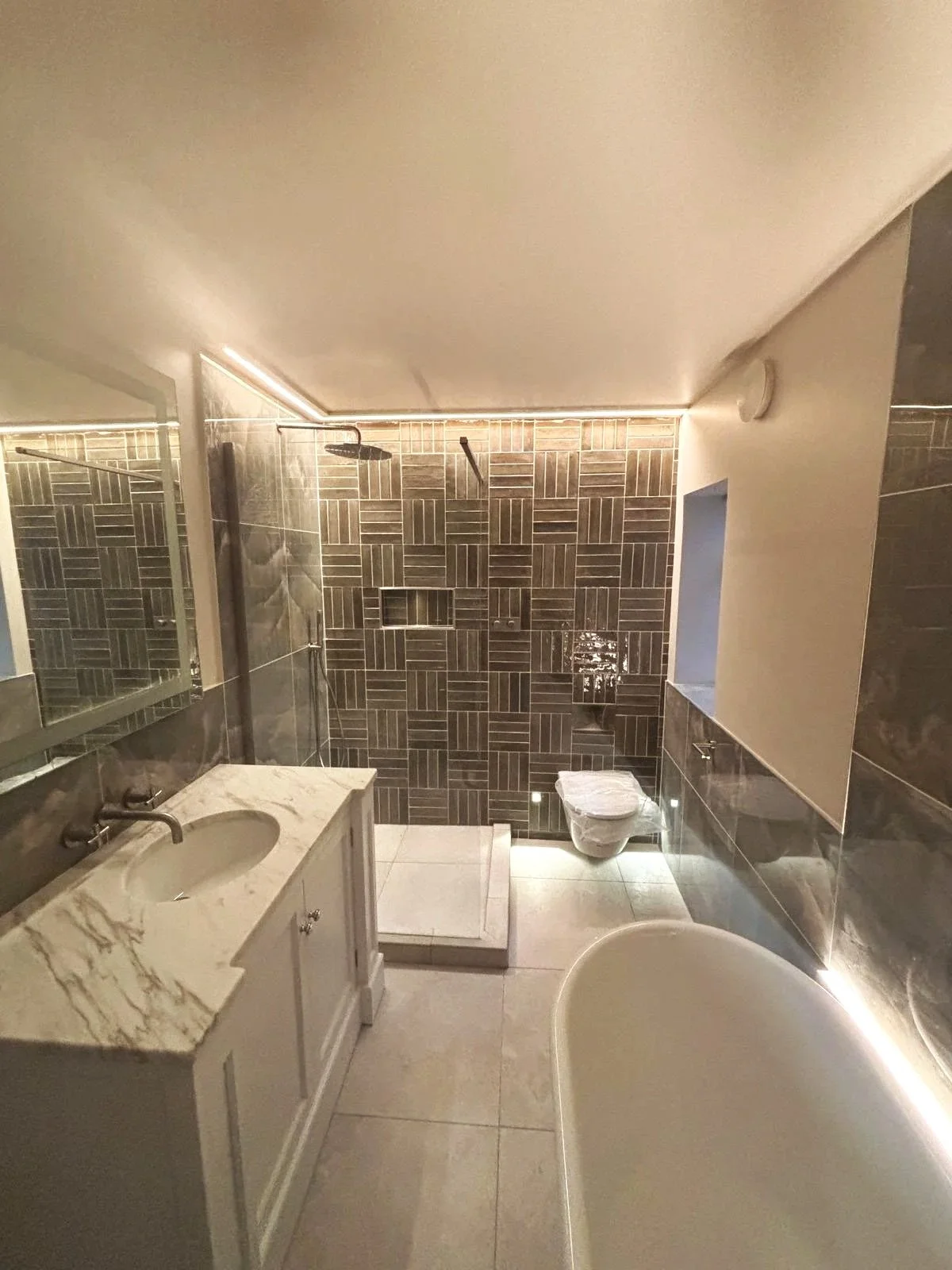 Modern bathroom with a marble vanity, a walk-in shower with dark tiles, a bathtub, and a toilet. Recessed lighting and a window are also visible.