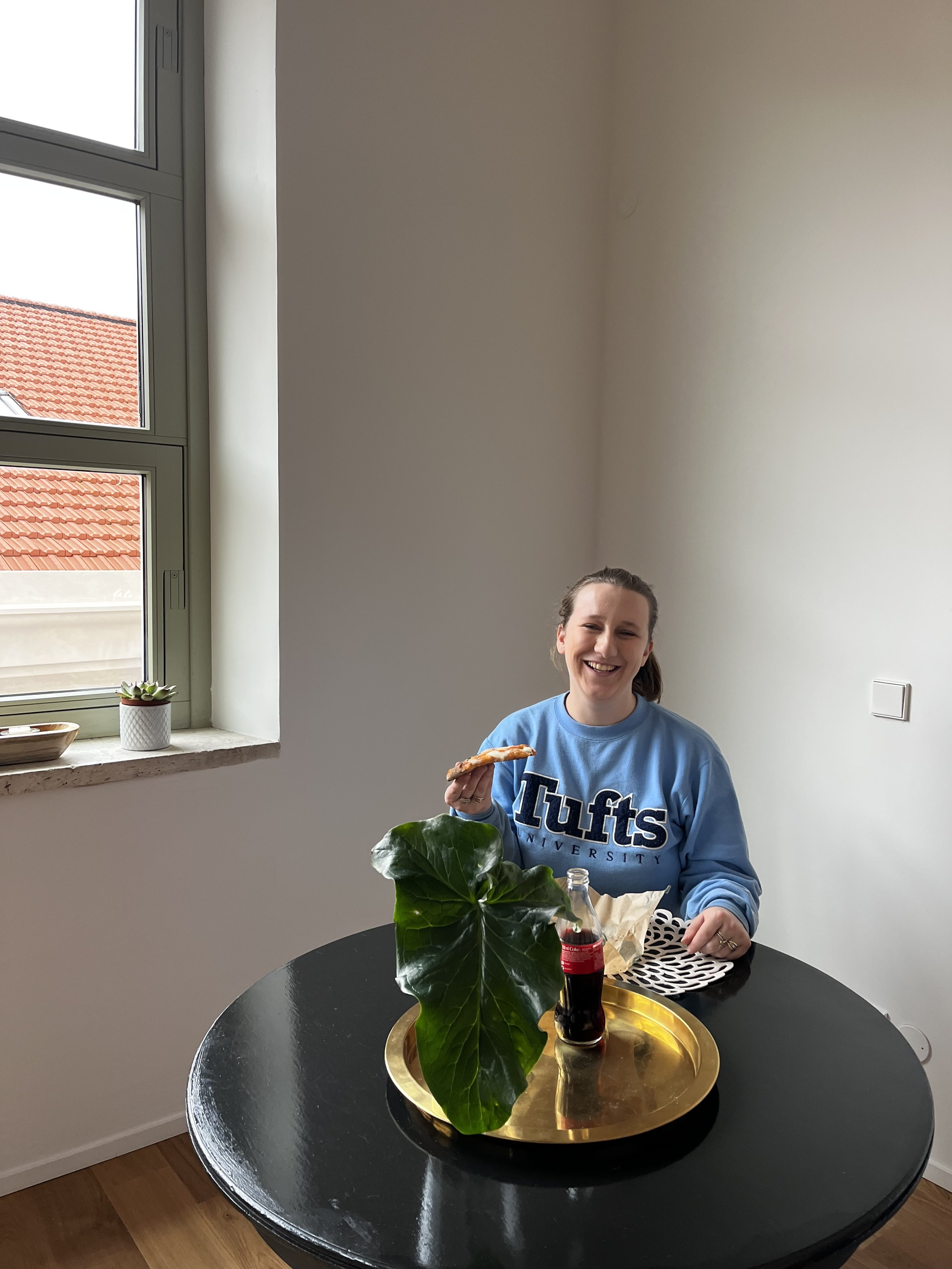 Person sitting at a round black table with a golden tray containing a large green leaf and a soda bottle; person is holding food and wearing a "Tufts University" sweatshirt.