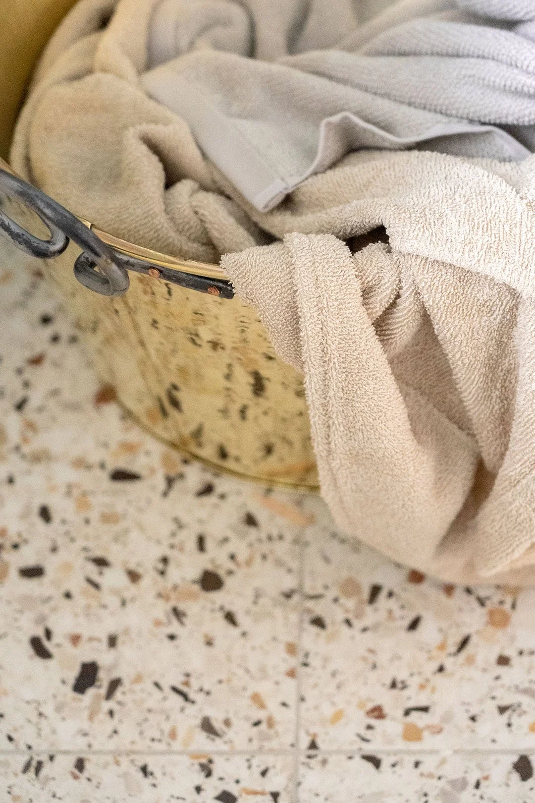 En messingkurv med beige og grå tøj, placeret på et terrazzo-flisegulv.