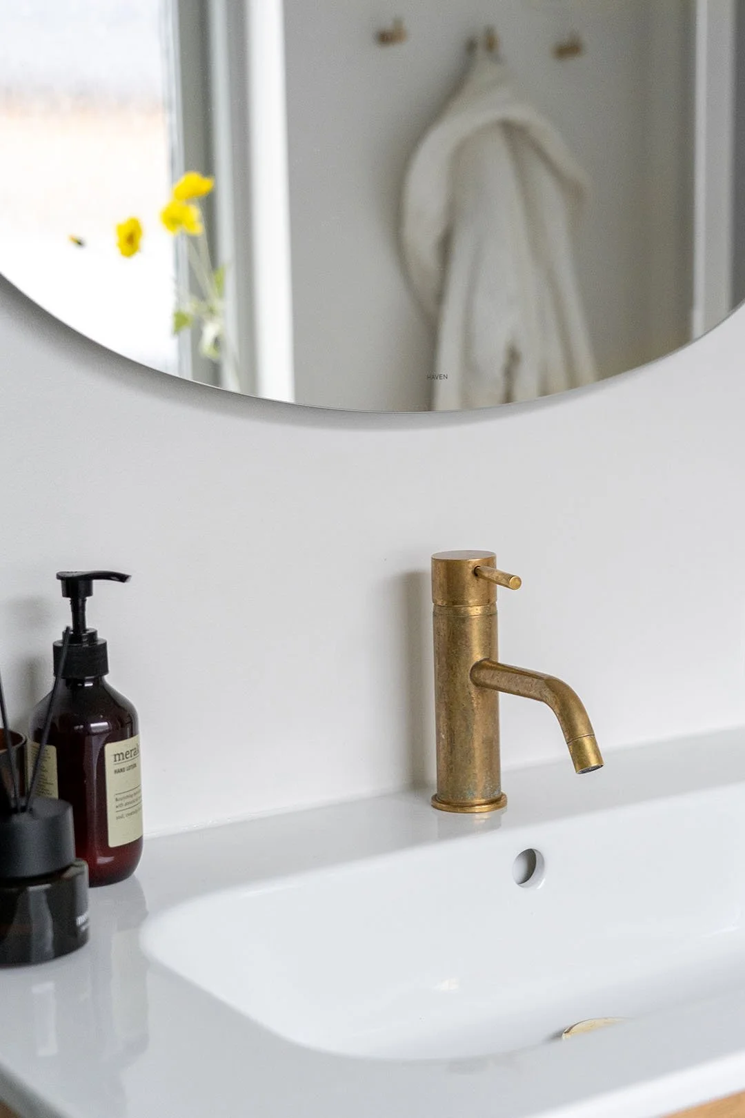 Hvid håndvask med messing armatur. Der er et rundt spejl over vasken og en hvid badekåbe eller håndklæde i baggrunden.