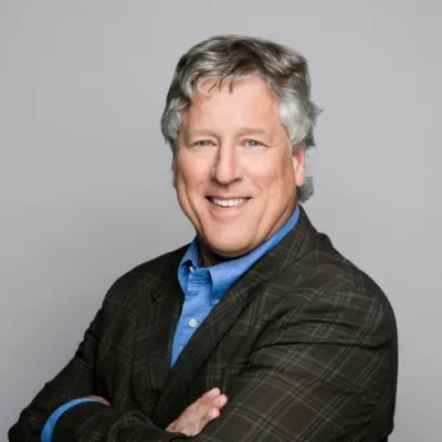 A smiling middle-aged man with gray hair, wearing a blue shirt and a dark plaid blazer, posing with arms crossed against a plain gray background.