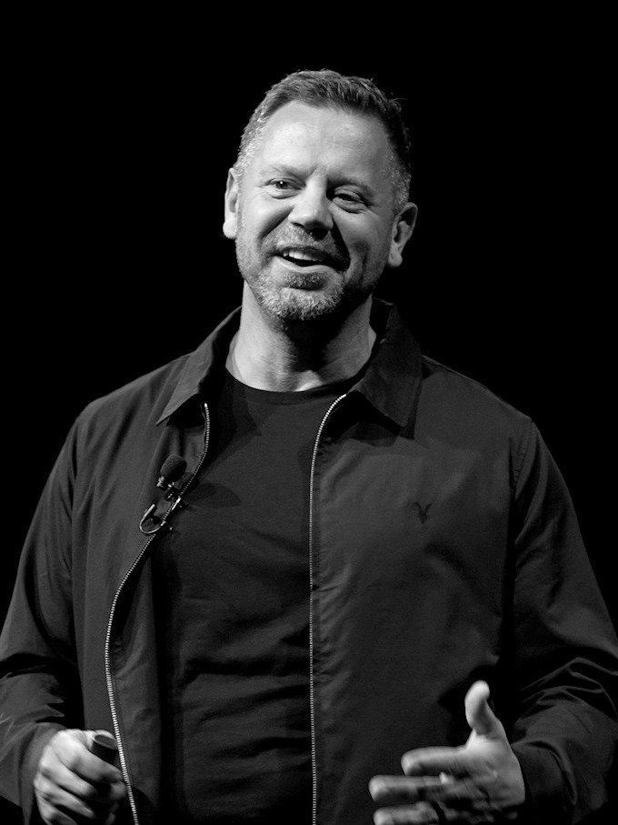 Black and white photo of a man smiling, wearing a black jacket and shirt, speaking into a microphone, standing against a plain dark background.