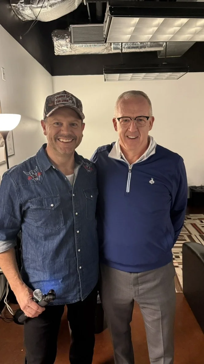 Two men smiling and standing close indoors. One man is wearing a denim shirt and baseball cap, holding a microphone. The other man is wearing glasses, a blue pullover with a zip, and gray trousers.