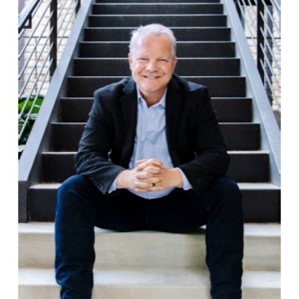 Older man with gray hair smiling, sitting on stairs outdoors, wearing a black blazer and light shirt.