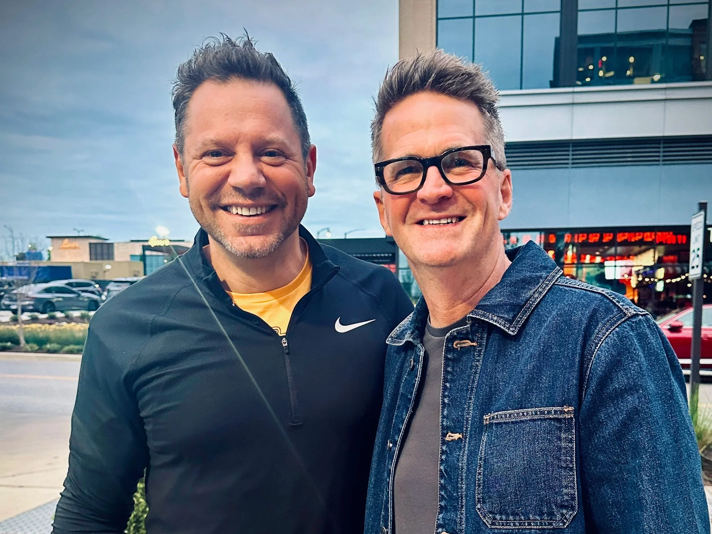 Two men are smiling and standing close together outdoors in a parking lot with a modern commercial building in the background. One man is wearing a black Nike jacket and the other a denim jacket with glasses.