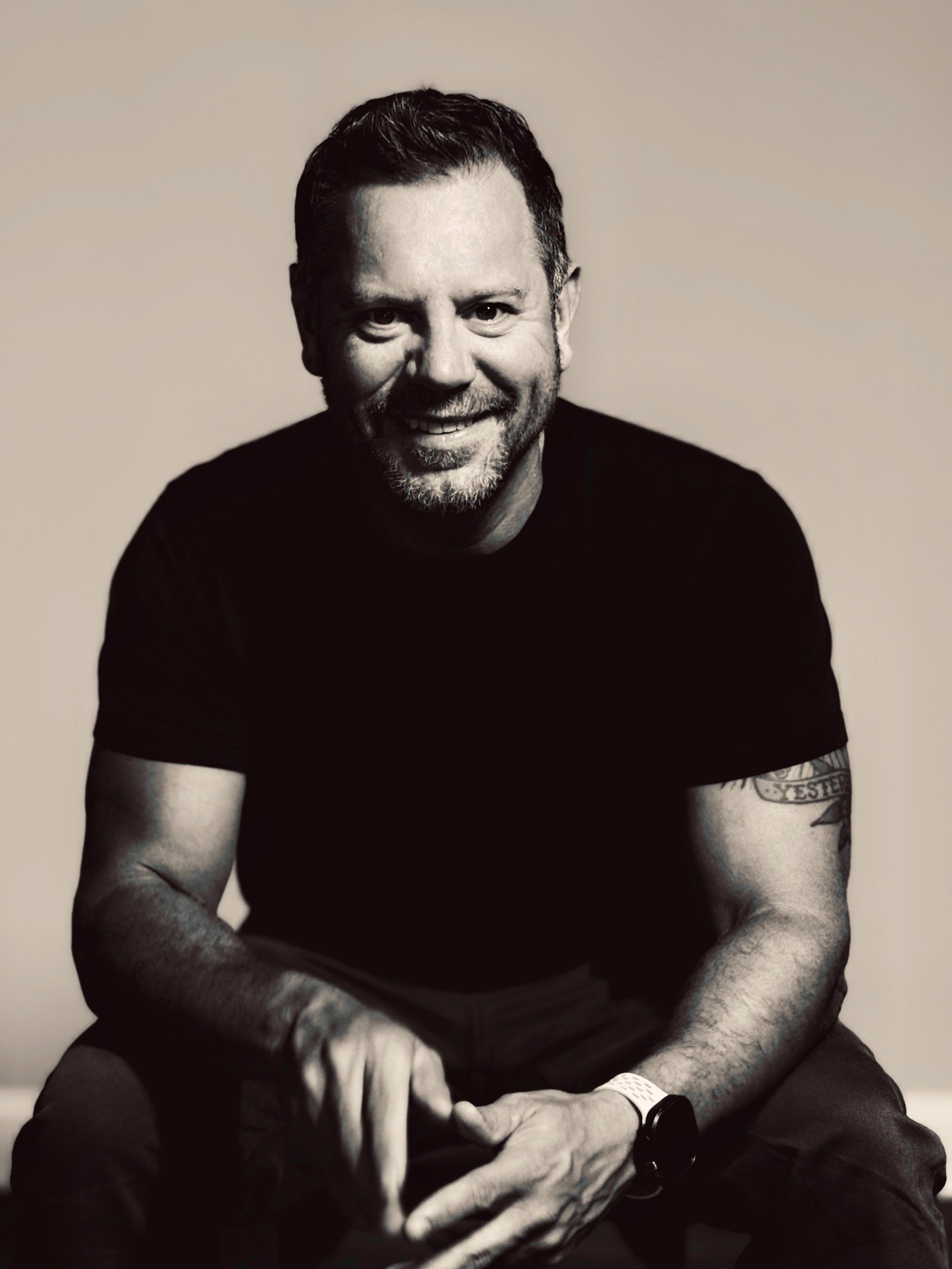 Black and white portrait of a smiling man with short, dark hair and facial hair, wearing a black t-shirt, sitting against a plain background.