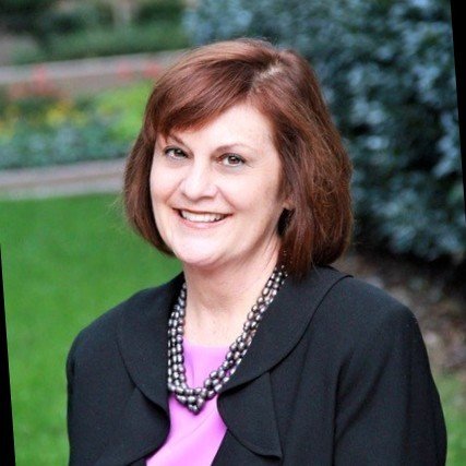 Smiling woman with shoulder-length reddish-brown hair, wearing a black blazer, purple top, and a pearl necklace, outdoors with greenery in the background.