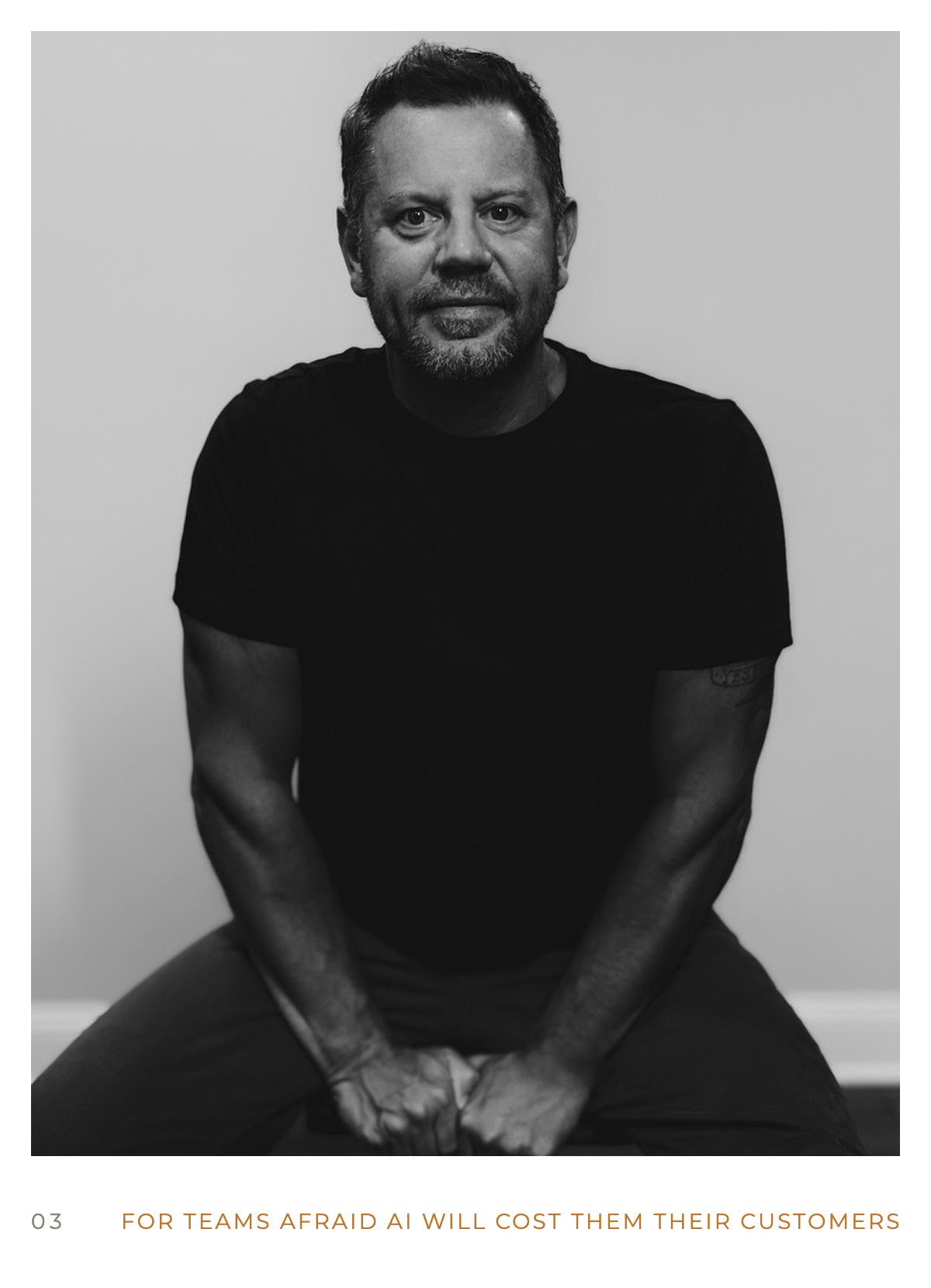Black and white photo of a man with short hair and a beard, wearing a black t-shirt, sitting with his arms resting on his legs against a plain wall.