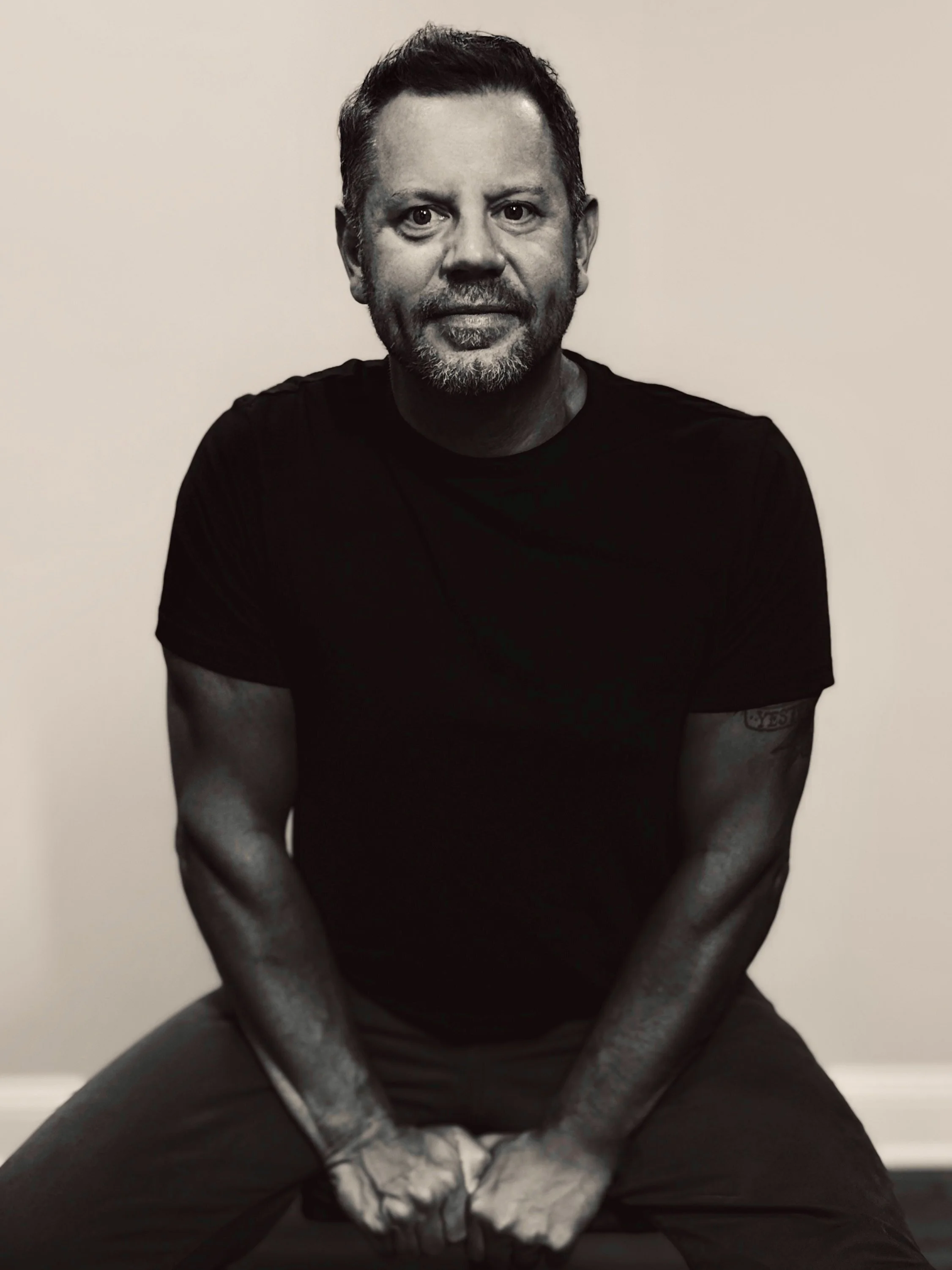 A man with dark hair, facial hair, and tattoos on his arms, sitting cross-legged against a plain white wall, wearing a black t-shirt.