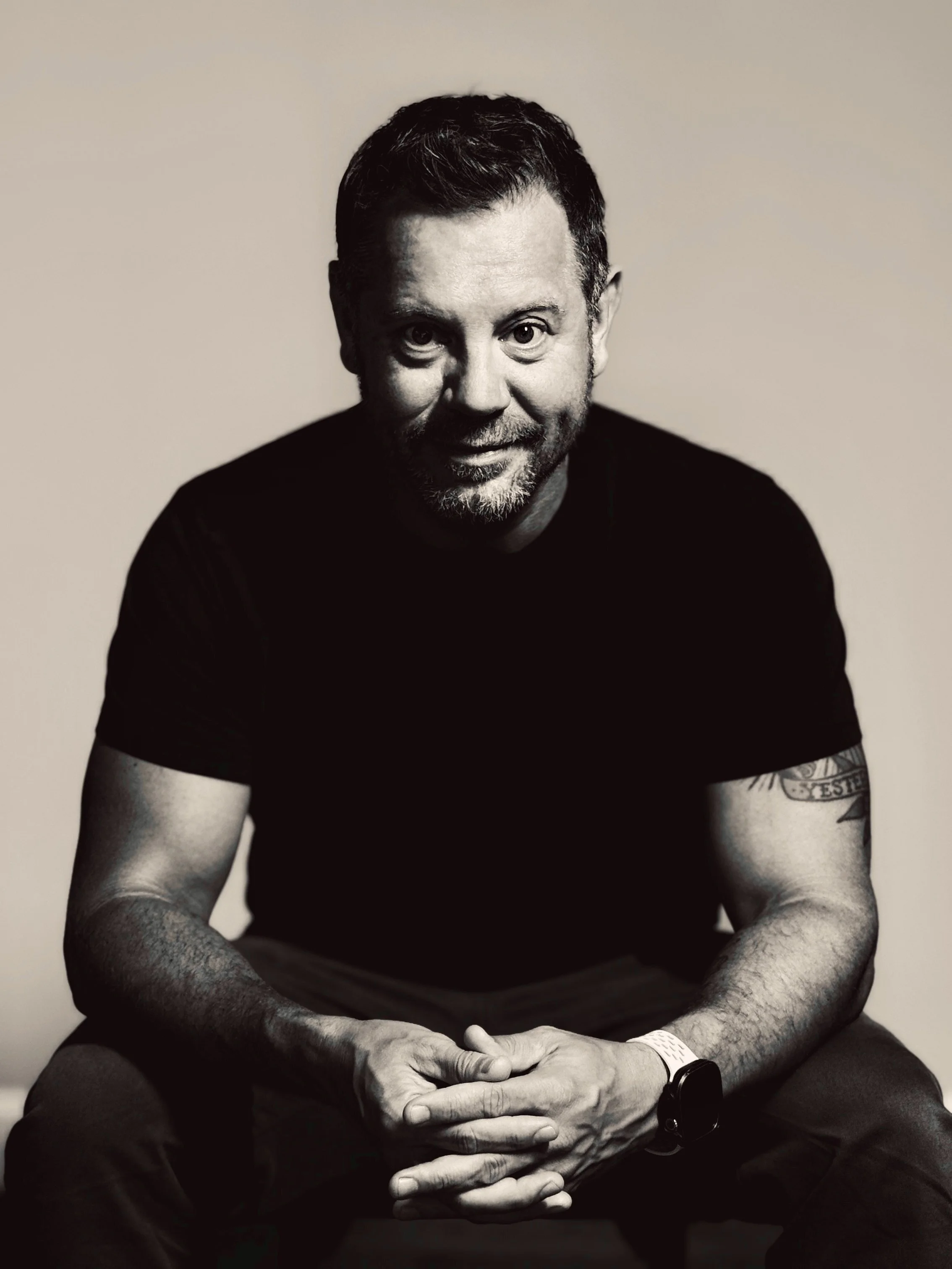 Black and white portrait of a man with short dark hair, a beard, and tattoos on his arms, sitting with his hands clasped in front of him, wearing a black shirt and a watch on his left wrist, against a plain light background.