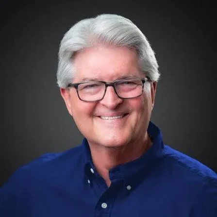 A smiling older man with gray hair and glasses, wearing a blue collared shirt against a dark background.
