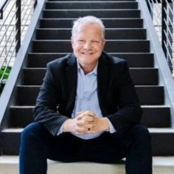 An elderly man with white hair, wearing a black suit and light blue shirt, sitting on outdoor stairs and smiling.