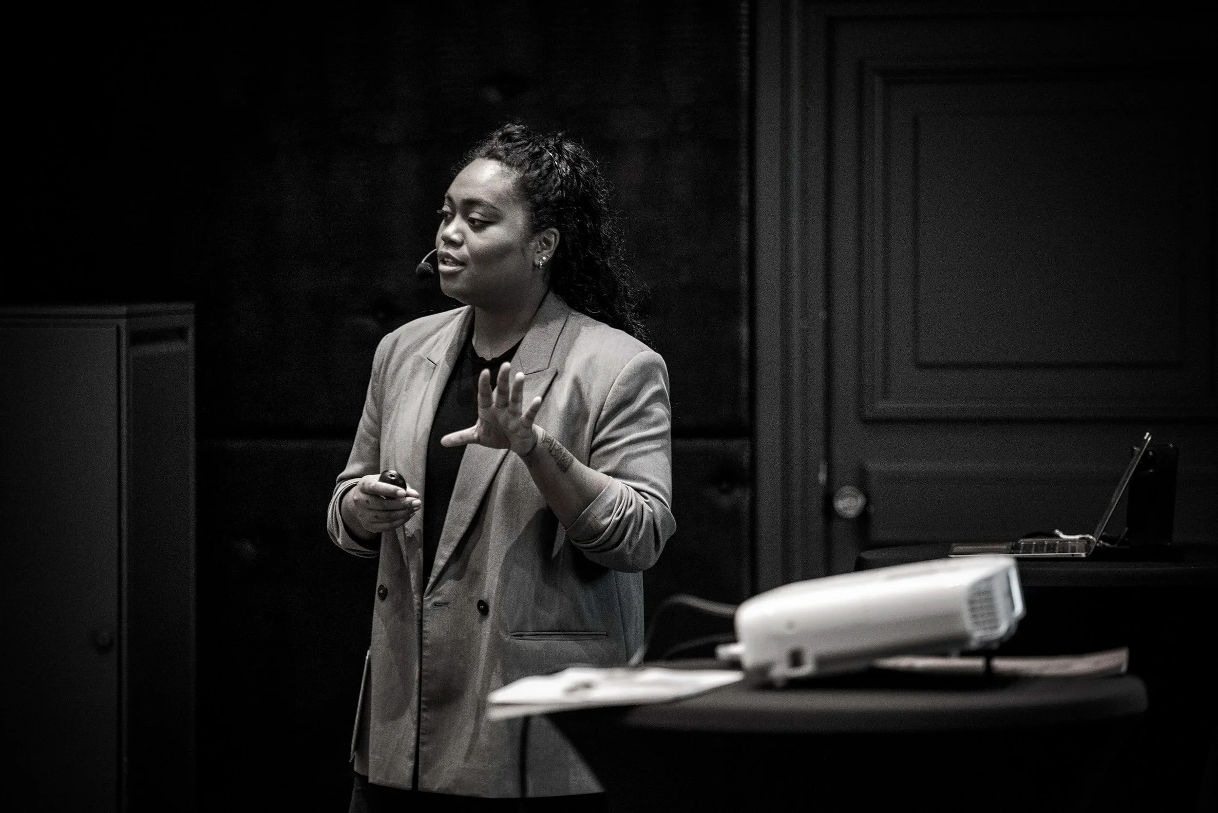 Femme donnant une présentation en noir et blanc, portant un blazer, avec un microphone dans l'oreille, dans une pièce sombre.