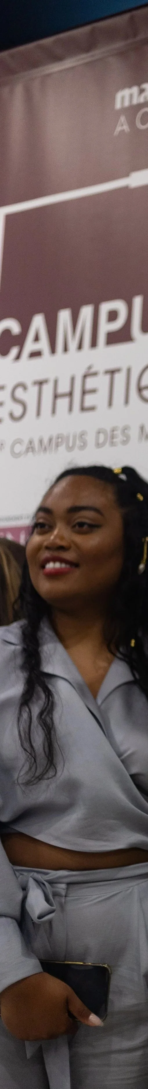 Une femme souriante avec des cheveux bouclés noirs portant une tenue grise se trouve devant un panneau mentionnant un campus d'esthétique.