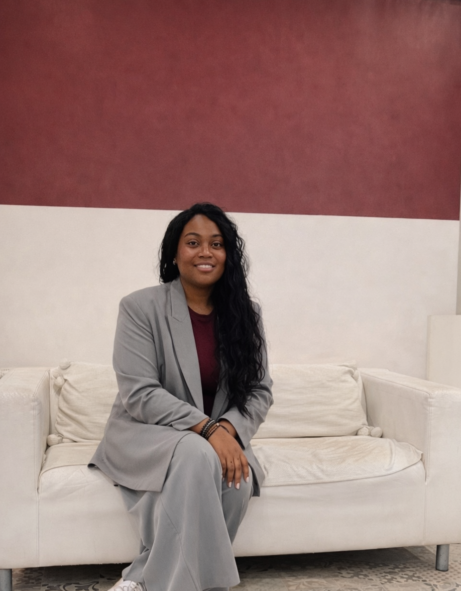 Femme assise sur un canapé blanc, portant un costume gris et un haut bordeaux, avec des cheveux longs et bouclés, dans un salon.