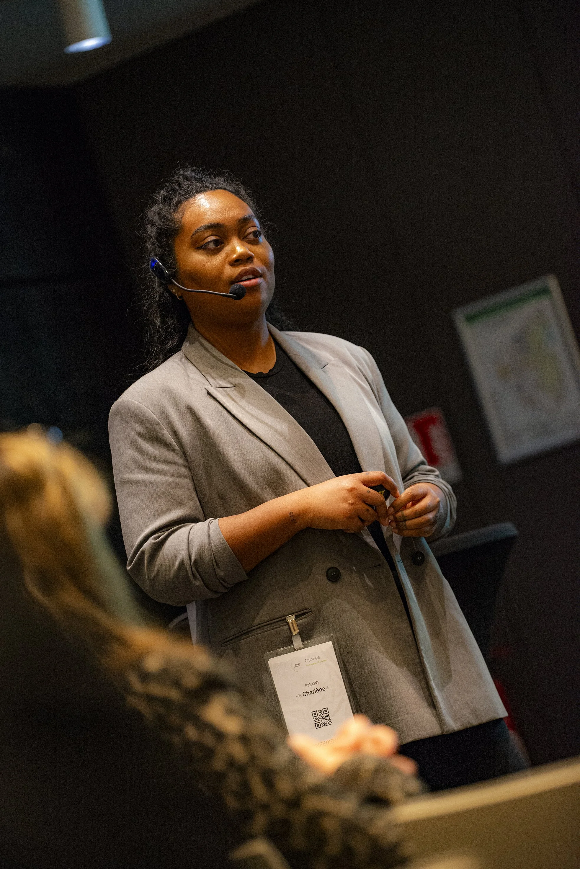 Une femme noire portant un blazer gris et une boucle d'oreille en argent parle lors d'une présentation, portant un casque avec microphone, dans une salle sombre.