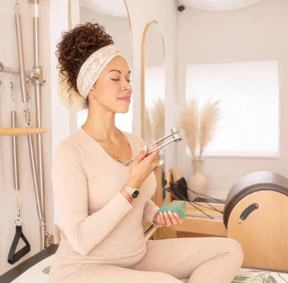 A woman practicing meditation or yoga in a bright, cozy room, sitting cross-legged with eyes closed, holding a green card in her hand. The room features soft lighting, neutral decor, and a large window with white blinds.