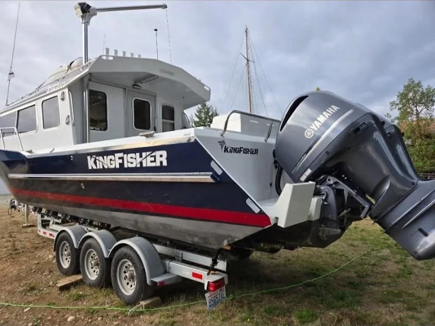 A KingFisher fishing boat on a triple-axle trailer, equipped with a Yamaha outboard motor, parked on grass with a cloudy sky and trees in the background.