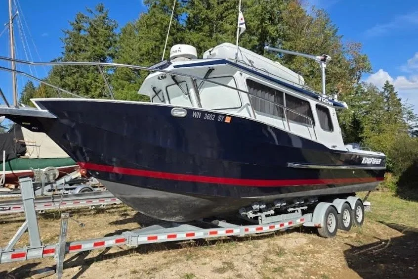 A large blue and white motorboat on a dual-axle trailer, parked on grass with trees in the background under a blue sky.
