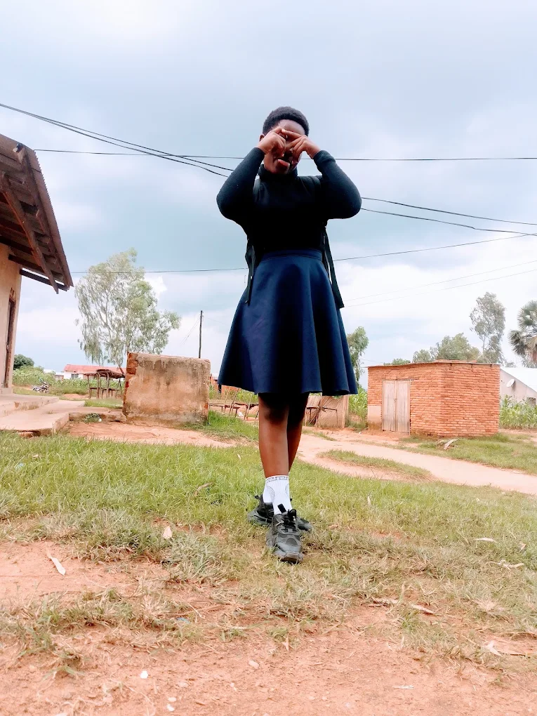 A young girl in a school uniform standing outdoors on a grassy area, covering her eyes with her hands, with houses and trees in the background under a cloudy sky.