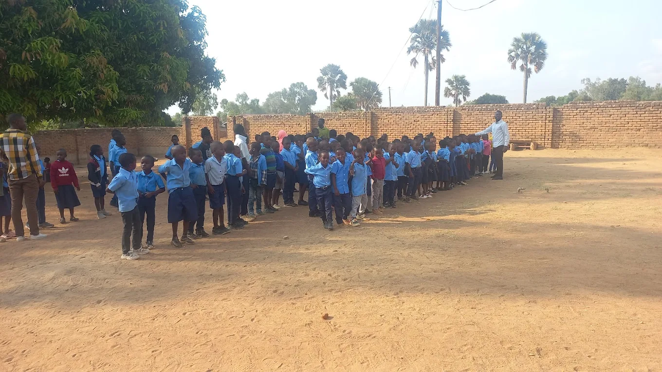 Group of school children standing in line outdoors on a dirt field, with teachers supervising. Many children are wearing blue school uniforms. New Palm Africa Huckleberry Fox Foundation