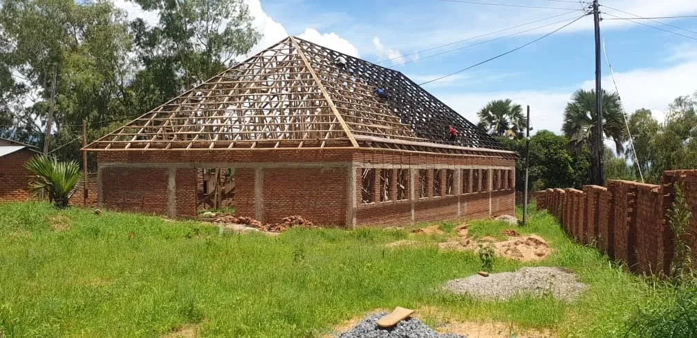 Brick building under construction with wooden roof frame, surrounded by grass and trees.