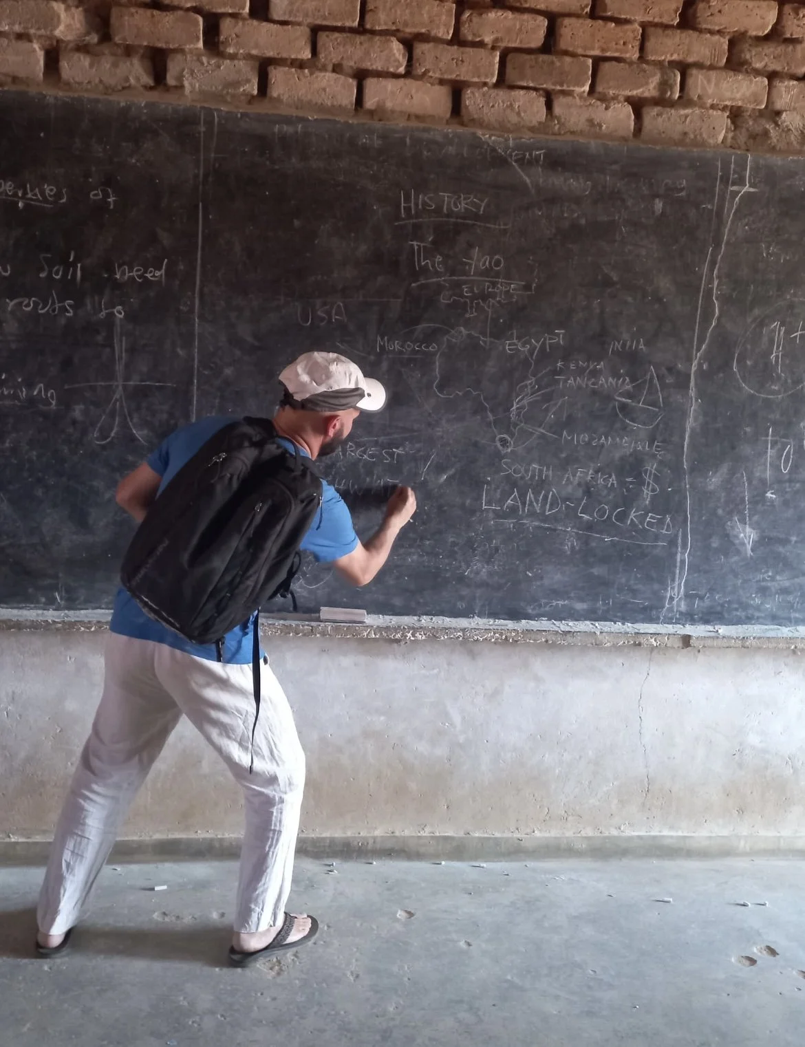 A man wearing a white hat and backpack writing on a blackboard with chalk, which contains a map of Africa, countries, and notes about history.