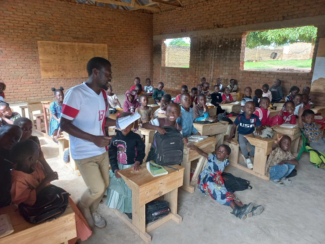 A classroom filled with young students seated at wooden desks, with a male teacher standing among them holding a notebook, in a brick-walled school house with open windows. New Palm Africa Huckleberry Fox Foundation