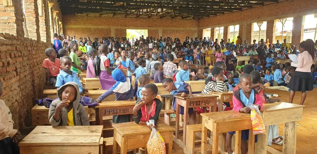 A large classroom filled with many children, sitting at wooden desks. The classroom has brick walls and an unfinished ceiling, with sunlight coming through the windows.